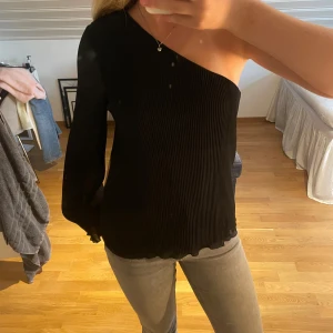 Svart ribbad oneshoulder topp - Säljer en svart ribbad topp med oneshoulder-design. Toppen har lång ärm på ena sidan och är ärmlös på andra, vilket ger en cool asymmetrisk look. Materialet är stretchigt och sitter snyggt på kroppen. Perfekt för dig som vill sticka ut med en trendig stil.