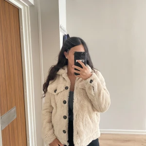 Beige teddyjacka från Viva Couture - Säljer en fluffig och supermjuk teddyjacka från Viva Couture i beige färg. Jackan har stora svarta knappar framtill, två bröstfickor med lock och långärmade ärmar. Perfekt för dig som gillar mysiga och trendiga jackor med oversized känsla.