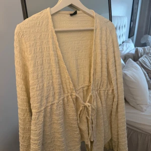 Beige omlottkofta från Gina Tricot - Superfin beige omlottkofta från Gina Tricot i storlek M. Koftan har ett ribbat, strukturerat tyg och knytband framtill. Långärmad och med djup v-ringning, perfekt att slänga på över en topp. Snyggt flowy material som känns mjukt mot huden.