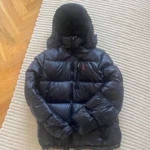 Svart Ralph Lauren Jacka - Jag säljer denna snygga och trendiga dunjackan från Ralph Lauren. Storleken är L men sitter även snyggt om du bär M. Riktigt bekväm och varm, den perfekta vinter jackan! Den har även innefickor och samtliga fickor har dragkedja. Luvan är även avtagbar med blixtlås vilket möjliggör stil variation! Säljer på grund av att jag nästan aldrig använt den. Skriv för eventuella frågor/fler bilder. Köpt på Ströms, nypris ca 4000kr—pris kan diskuteras!