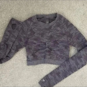 Lila camo croppad topp från Gymshark - Säljer en croppad långärmad topp från Gymshark i lila camouflage-mönster. Toppen har ribbad halsringning, tumhål i ärmsluten och snygg rynkdetalj framtill. Perfekt för träning eller chill dagar.