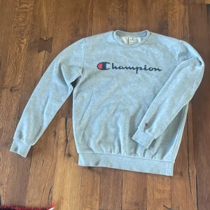 Grå Champion sweatshirt i bomull - Grå sweatshirt från Champion i storlek S med klassisk logga tryckt över bröstet. Tröjan har rund hals, långa ärmar och ribbade muddar vid ärmslut och nederkant. Perfekt för chill dagar och enkel att matcha med jeans eller joggers.