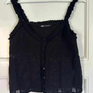 Spetsig linnetopp från Zara - Svart linnetopp från Zara med spetsdetaljer och broderade blommor. Toppen har v-ringning, knappar framtill och fransiga axelband. Croppad passform som ger en cool vibe.🍒🍒🌸