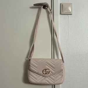 Beige quiltad axelväska med gulddetalj - Snygg beige axelväska i quiltad design med guldfärgad GG-detalj framtill. Väskan har justerbar axelrem, dragkedjeficka på baksidan och flera fack inuti. Perfekt storlek för det viktigaste och ger en clean look till din outfit.