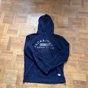 Mörkblå hoodie från Jack & Jones - Säljer en mörkblå hoodie från Jack & Jones med tryck på bröstet. Tröjan har huva och en stor magficka framtill. Perfekt för dig som gillar en avslappnad och sportig stil. Klassisk passform och mjukt material.