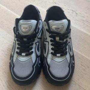 D i o r sneakers CD - Snygga  B30 sneakers med svart och silverfärgad ovandel, reflekterande detaljer och tydlig CD-logga på hälen. Skorna har meshpaneler, svarta snören och en chunky sula. Perfekta för dig som vill sticka ut med exklusiva och trendiga sneakers.