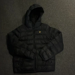 Svart pufferjacka från Lyle & Scott - Svart pufferjacka från Lyle & Scott med huva och dragkedja. Jackan har det klassiska gula fågel-loggan på bröstet och är vadderad för extra värme. Perfekt för kalla dagar. Om du beställer från bring kan jag leverera paketet mycket snabbt.de är storlek 12-13 men sitter som 14 priset kan diskuteras.