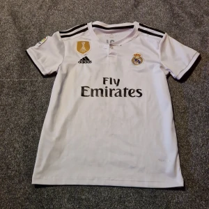 Real Madrid Bale matchtröja + shorts - Säljer en vit Real Madrid matchtröja med Bale 11 på ryggen och tillhörande shorts. Tröjan har svarta detaljer, klubbmärke, Adidas-logga och Fly Emirates-tryck. Korta ärmar och klassiska svarta ränder på axlar och shorts. Materialet är lätt och andas, perfekt för sport..