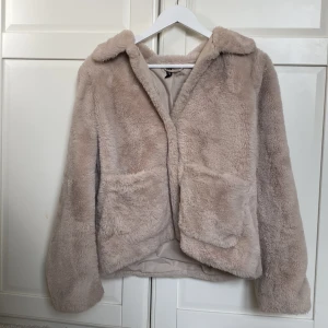 Beige pälsjacka från H&M Divided - Supermjuk och fluffig beige pälsjacka från H&M Divided i storlek S. Jackan har två stora fickor framtill, bred krage och stängs med knappar. Perfekt för dig som gillar en cozy och trendig look under kyliga dagar.