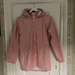 Rosa regnjacka från Everest med huva - Säljer en snygg rosa regnjacka från Everest i storlek 164. Jackan har huva, vita tryckknappar, fickor framtill och reflexdetaljer på ryggen. Perfekt för regniga dagar och enkel att matcha med din stil.