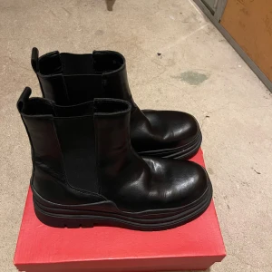 Svarta chelsea boots från XIT - Chunky svarta chelsea boots från XIT i skinn med elastiska paneler på sidorna och grov platt sula. Snygg minimalistisk design med rund tå och dragflik bak. Perfekta för dig som gillar en edgy och stilren look.
