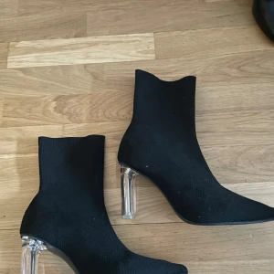 Svarta stövletter med transparent klack - Trendiga svarta stövletter i stickat material med spetsig tå och hög transparent klack. Snygg och modern siluett som ger en edgy vibe till outfiten. Perfekta för dig som vill sticka ut med dina skor.