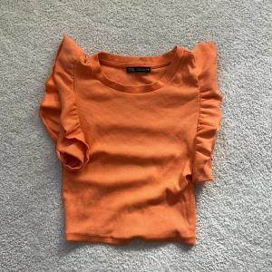 Orange ribbad topp med volangärmar Zara - Superfin orange ribbad topp från Zara med korta volangärmar. Toppen har rund halsringning och är i ett stretchigt material som sitter snyggt på kroppen. Perfekt för dig som vill ha en färgklick i garderoben!