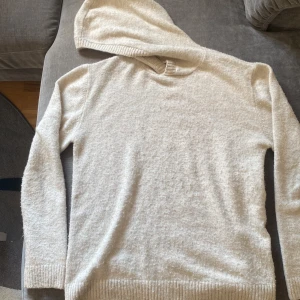 Beige stickad hoodie från H&M - Mysig beige stickad hoodie från H&M i storlek 158/164. Tröjan har lång ärm, ribbade muddar och en klassisk huva. Perfekt för kyliga dagar när du vill ha något både bekvämt och stilrent. Tyvärr finns det 1 litet hål i båda axelstyngnen på tröjan
