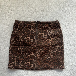 Leopardmönstrad kortkjol med dragkedja - Snygg kortkjol med leopardmönster i bruna och svarta toner. Kjolen har en cool dragkedja framtill med ringdetalj och är figurnära i modellen. Perfekt för dig som vill sticka ut och addera lite edge till din outfit.