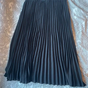 Svart plisserad långkjol - Snygg svart plisserad långkjol med bred midja och elegant fall. Kjolen har klassiska veck som ger en stilren och trendig look. Perfekt att matcha med en croppad topp eller stickad tröja för en cool outfit.