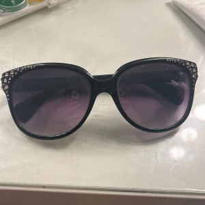 Svarta solglasögon med strass Montini - Stora svarta solglasögon från Montini med glittriga strasstenar på bågens ovansida. Snygg oversized look och mörka glas. Perfekta för att ge en extra touch till din outfit.