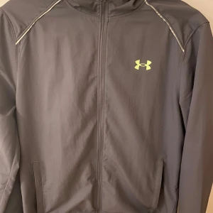Svart vindjacka från Under Armour - Svart vindjacka från Under Armour med huva och dragkedja framtill. Jackan har neongult logotryck på bröstet och stor Under Armour-text på ryggen. Tillverkad i lätt polyester, perfekt för träning eller när du vill ha en sportig look. Storlek S och även i bra skick