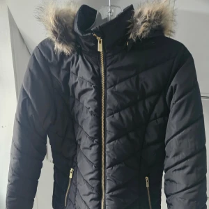 Svart quiltad pufferjacka med fuskpäls strl 164 - Svart pufferjacka med snyggt quiltat mönster och guldiga dragkedjor. Jackan har huva med fuskpäls och två fickor framtill. Perfekt för kalla dagar när du vill hålla stilen och värmen.