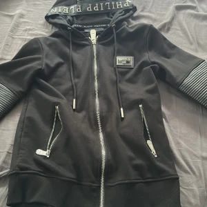 Philipp Plein svart hoodiejacka - Svart hoodiejacka från Philipp Plein med dragkedja, ribbade detaljer på ärmarna och två snedställda fickor med dragkedja. Stor dödskalle i blått och silver på ryggen, samt logga på huvan och bröstet. Perfekt för dig som gillar edgy streetstyle.