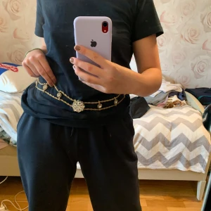 Glittrigt guldigt kedjebälte med strass - Säljer ett unikt kedjebälte i guld med glittrande strassdetaljer och en stor, rund strassdekoration framtill. Bältet har tvinnade kedjor och små strassprydda cirklar längs hela längden. Perfekt för att ge din outfit extra bling och statement.