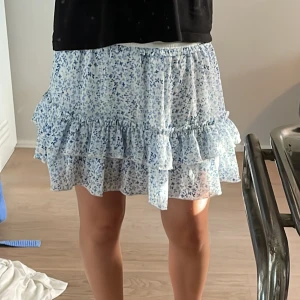 Blommig volangkjol i blått och vitt - Supersöt kort kjol med blått blommönster på vit botten. Kjolen har volangdetaljer och resår i midjan för en bekväm passform. Perfekt för varma dagar och ger en fräsch och somrig vibe. Lappar är avklippta men tror den är köpt på newyorker.