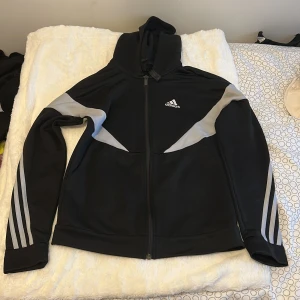 Svart Adidas hoodie med grå detaljer - Svart hoodie från Adidas med gråa paneler över axlar och armar samt klassiska tre ränder längs ärmarna. Dragkedja framtill, huva och Adidas-logga på bröstet. Perfekt för träning eller chill. Materialet är mjukt och sportigt, troligen polyester.