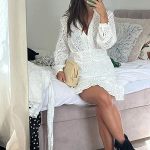 Vit kort klänning med broderade detaljer - Superfin vit kort klänning med broderade hålmönster och volangkant nertill. Klänningen har lång ärm, markerad midja och v-ringning med knappar framtill. Perfekt för sommarens alla tillfällen och ger en fräsch och romantisk vibe.