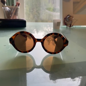Runda leopardmönstrade solglasögon NA-KD - Snygga runda solglasögon från NA-KD med leopardmönstrad båge i bruna och svarta toner. Glasen är bruna och bågarna är i plast med en cool retrovibe. Perfekta för att sticka ut och ge din look en extra edge . De är väldigt unika och snygga! Leopardmönster vilket är coolt
