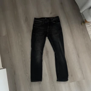 Svarta jeans från Hugo Boss - W30 L32. Snygga svarta jeans från Hugo Boss med klassisk femficksdesign och diskret tvättade detaljer. Jeansen har normal passform och raka ben, perfekt för en stilren look. Mjukt och bekvämt material i bomullsblandning. Läderpatch med BOSS-logga bak i midjan.