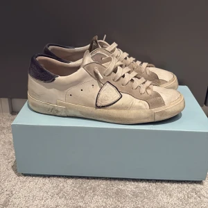 Philippe Model vita sneakers i skinn - Snygga vita sneakers från Philippe Model med grå mockadetaljer och mörkblå häl. Klassisk låg modell med snörning, rund tå och platt sula. Skorna har märkets ikoniska sköld på sidan och är tillverkade i skinn och mocka för en lyxig känsla. Har varit välldigt sparsamt använda. De är i storlek 41 men passar 42. Man får även med dustbag, tag och kartong i fint skick.