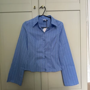 Blå randig figursydd skjorta från princess polly - Snygg blå skjorta från princess polly med vita tunna ränder, klassisk krage och raka långa vida ärmar. Skjortan har knäppning framtill med klädda knappar. Perfekt för dig som gillar en clean och fräsch look.