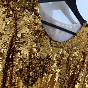 Guldig paljettklänning med svart knyt - Säljer en glittrig guldig kortklänning med paljetter och långärmade puffärmar. Klänningen har en rund halsringning och ett svart knytband i midjan som ger en snygg kontrast. Perfekt för dig som vill sticka ut och glänsa på festen!