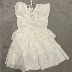 Vit kort klänning med volanger och snörning - Supersöt vit klänning från en marknad med broderade detaljer och volanger i flera lager. Smockad överdel och snörning fram ger en snygg passform. Perfekt för sommarens alla tillfällen