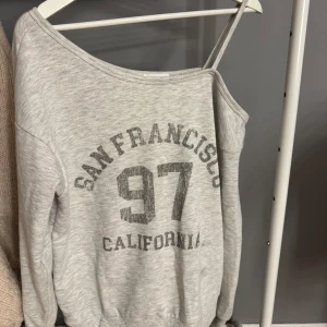 Grå offshoulder tröja Even&Odd - Grå sweatshirt från Even&Odd med tryck 'San Francisco 97 California' framtill. Tröjan har en offshoulder-design med en axelbandsdetalj och långa ärmar. Perfekt för en avslappnad och trendig look.