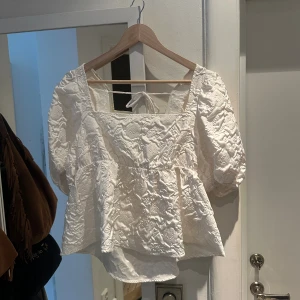 Vit blus med puffärmar från H&M - Superfin vit blus med broderade detaljer och puffärmar från H&M. Toppen har fyrkantig ringning, knytning i ryggen och lätt volang nedtill! Använd ca 1 gång så i väldigt fint skick. Justerbar i ryggen så passar även L🥰