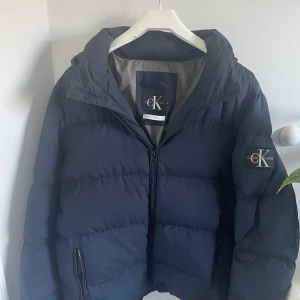 Calvin Klein jacka - Säljer en snygg mörkblå dunjacka från Calvin Klein Jeans. Jackan har huva, dragkedja framtill och två fickor med dragkedja. Stor CK-logga på ärmen ger en clean och modern look. Perfekt för kalla dagar när du vill vara både varm och stilren.