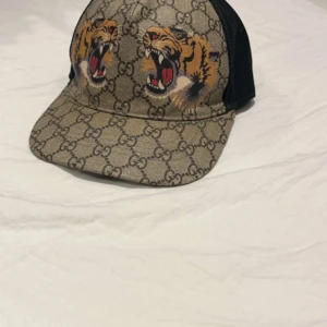 Gucci keps med tigertryck - Snygg keps från Gucci med klassiskt GG-mönster i beige och brunt, prydd med två coola tigerhuvuden framtill. Baksidan är i svart mesh för extra ventilation. Perfekt för dig som vill sticka ut med lyxig streetstyle.