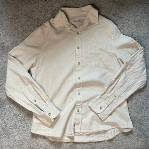 Beige linneskjorta från Dressmann - Snygg beige skjorta från Dressmann i linnemix. Klassisk krage, bröstficka och knäppning framtill. Långärmad modell med en avslappnad passform, perfekt för en stilren look. Skjortan är lätt och luftig, vilket gör den grym för varmare dagar.