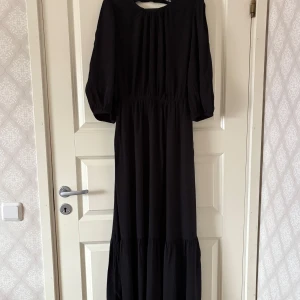 Svart långklänning med öppen rygg - Säljer en svart långklänning  med puffiga långärmar och snygg öppen rygg med knyt. Klänningen har rund halsringning och är tillverkad i ett mjukt, lätt polyestertyg som faller snyggt. Perfekt för dig som gillar stilrena och trendiga plagg.