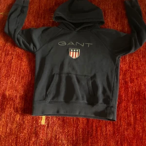 Svart hoodie från GANT med logga - Svart hoodie från GANT med stor broderad logga och sköld på bröstet. Klassisk känguruficka framtill och huva. Perfekt för dig som gillar en stilren och sportig look. Mjuk och skön bomullskvalitet.