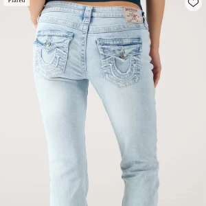 Ljusblå bootcut jeans True Religion - Säljer ett par ljusblå bootcut jeans från True Religion med ikoniska broderade bakfickor och silverfärgade knappar. Jeansen har låg midja och är tillverkade i klassisk denim med snygg tvätt. Perfekta för dig som gillar en avslappnad men trendig look.