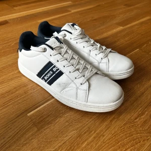 Vita sneakers från Björn Borg - Fräscha vita sneakers från Björn Borg med svarta detaljer och logga på sidan. Skorna har snörning, rund tå och är tillverkade i skinn med textilfoder. Perfekta för en clean och sportig look.