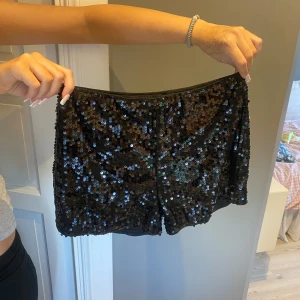 Svarta paljettshorts med loose fit - Glittriga svarta shorts täckta med paljetter som ger en riktigt festlig vibe. Modellen är loose fit och har en rak, enkel form med elastisk midja. Perfekta för att sticka ut och glänsa på nästa event. Materialet är syntet och känns lätt mot huden.