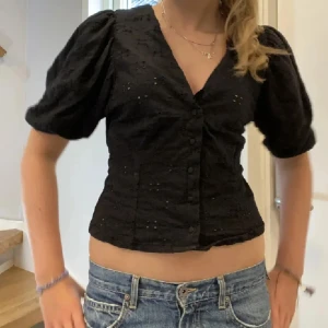Svart blus med puffärm och knappar - Supersnygg svart blus med puffiga korta ärmar och v-ringning. Blusen har knappar framtill och är sydd i ett lätt bomullstyg med broderade håldetaljer. Sitter figurnära och är perfekt till jeans eller kjol.