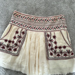 Broderad kortkjol i bomull - Superfin kortkjol i ljusbeige bomull med broderade blommor i rött och svart. Kjolen har bred midja med broderade detaljer och dragkedja i sidan. Knappt använd. 