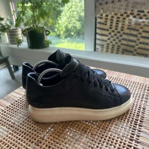 Alexander McQueen svarta sneakers - Svarta sneakers från Alexander McQueen med chunky vit platt sula och klassisk snörning. Skorna är i skinn och har rund tå samt diskret logga bak på hälen. Perfekta för dig som gillar stilrena och trendiga sneakers med extra höjd.