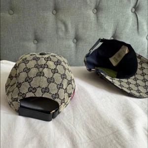 Gucci keps med GG-mönster och röd-blå detalj - Säljer en beige keps från Gucci med klassiskt GG-mönster i mörkgrått. Kepsen har en böjd skärm och justerbar rem baktill i svart. På sidan finns en snygg röd-blå rand som ger extra lyxig vibe. Materialet är bomullscanvas och insidan är fodrad i mörkblått tyg.
