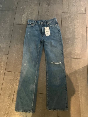Blå raka jeans med slitning - Säljer ett par blå raka jeans med slitning på ena benet och klassisk femficksmodell. Jeansen har hög midja och är tillverkade i denim med en snygg tvätt. Perfekta för en avslappnad och trendig look.