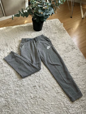 Gråa mjukisbyxor från Nike - Säljer ett par gråa mjukisbyxor från Nike med klassisk logga på vänster ben. Byxorna har resår i midjan, två sidofickor och muddar vid bensluten. Perfekta för chill dagar eller när du vill vara bekväm. Materialet är mjukt och skönt. Om du har några frågor skriv till mig, svarar så snabbt jag kan!🙌